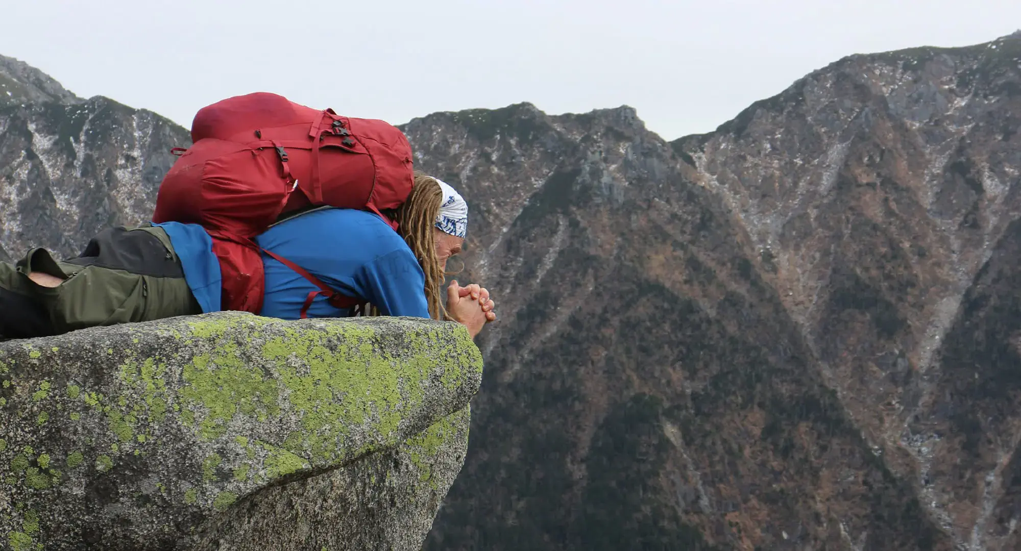 A woman with a red backpack lying down and peering over the edge of a cliff