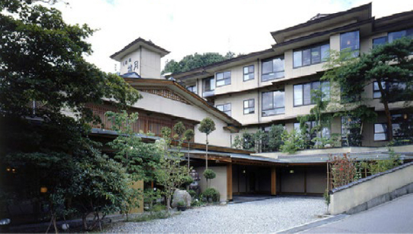 The exterior of a traditional Japanese ryokan, featuring a wooden structure, lush greenery, and a welcoming entrance with classic Japanese architecture.