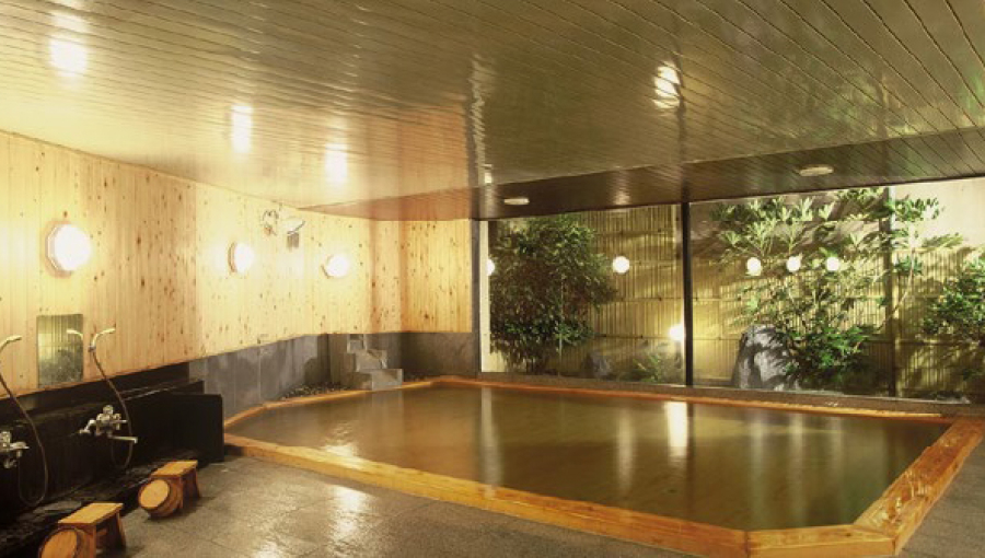 A spacious indoor onsen bath with warm lighting, wooden paneling, and a relaxing atmosphere, offering a traditional Japanese hot spring experience.