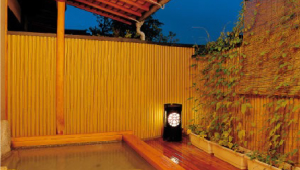 An outdoor onsen bath surrounded by a bamboo fence and greenery, illuminated by a warm lantern, creating a tranquil and intimate ambiance.