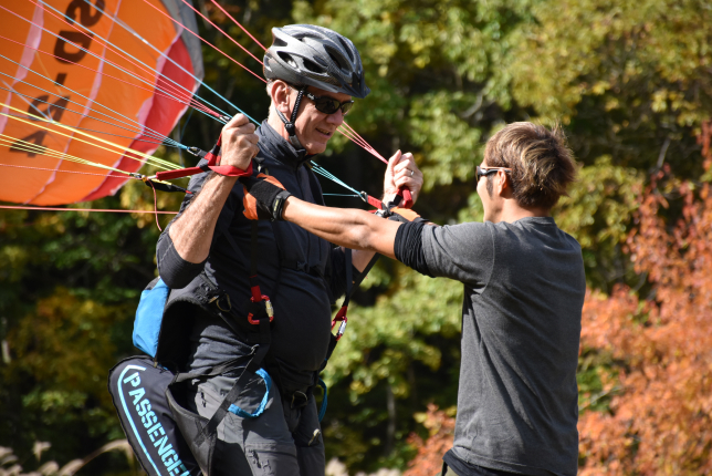 A paragliding instructor assisting an older man in a black outfit and helmet, preparing for a tandem flight. The parachute strings are secured as the man gets ready for takeoff.