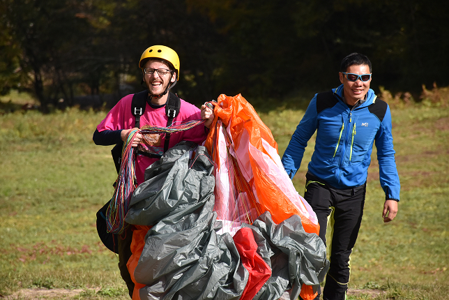 A smiling man wearing a yellow helmet and pink shirt, carrying a parachute canopy after landing. Another person in sunglasses and a blue jacket walks beside him, both looking excited.