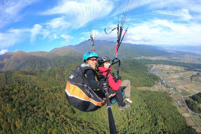 A tandem paragliding experience high above a scenic valley. The pilot and passenger, wearing helmets and warm clothing, capture the breathtaking view with a selfie stick.
