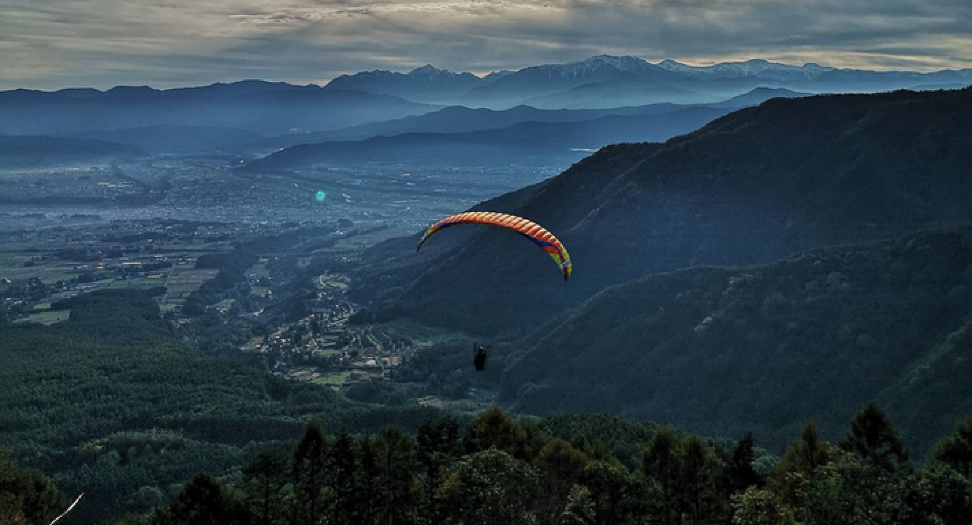 A paraglider soaring over a vast mountain range with a valley below. The setting sun casts a golden hue over the landscape, creating a stunning aerial perspective.