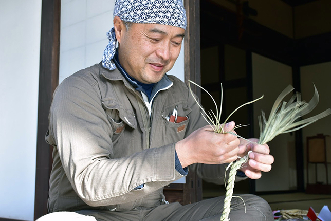A man wearing a blue-patterned headscarf and a brown jumpsuit is weaving straw into a rope while smiling, sitting on the wooden floor of a traditional Japanese house.