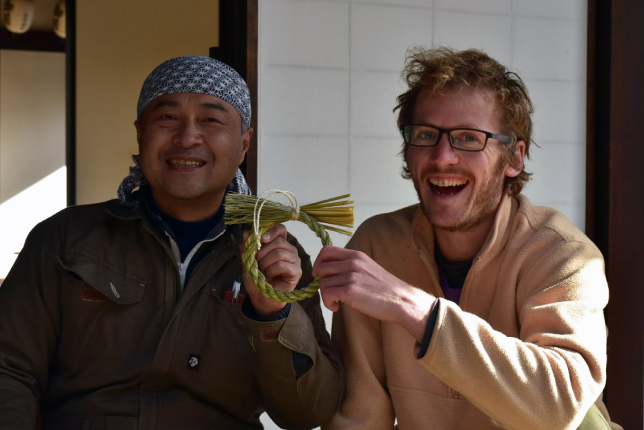 Two men sitting outside a traditional Japanese house, smiling while holding a handmade straw ornament together.