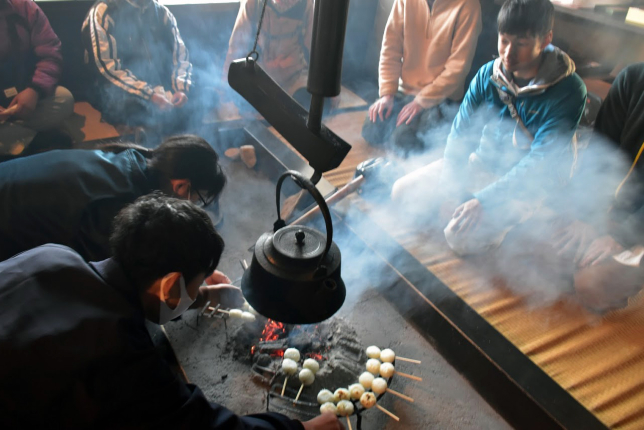 A group of people gathered around an irori (traditional Japanese sunken hearth) roasting skewered dumplings over a charcoal fire in a dimly lit room.