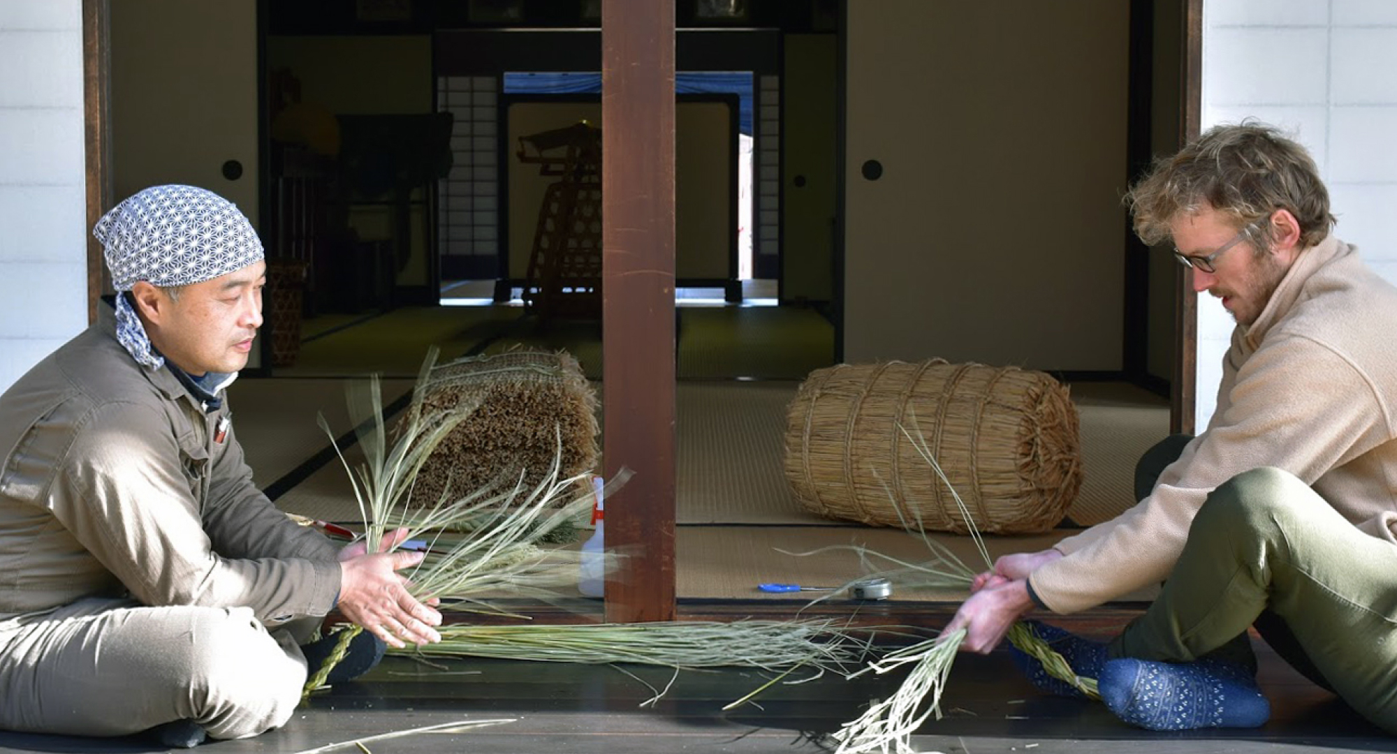 A Japanese artisan and a foreign participant sitting on the wooden veranda of a traditional house, weaving straw together with baskets and tools nearby.