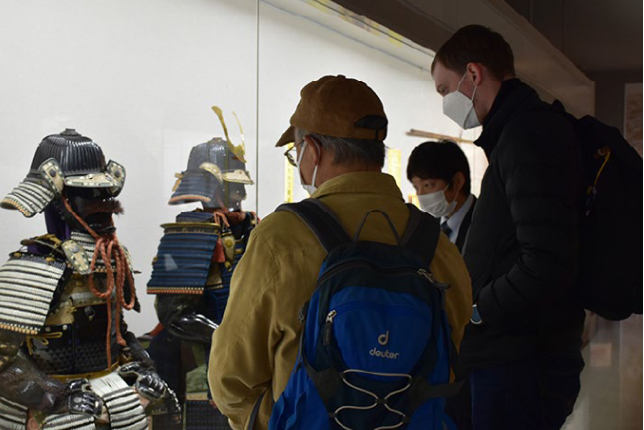 A group of visitors, including an older man in a cap and a younger man with a blue backpack, observing a display of traditional samurai armor inside a museum.