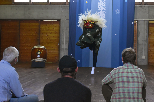 A traditional Japanese lion dance performance in a wooden hall, with an audience watching in the foreground.