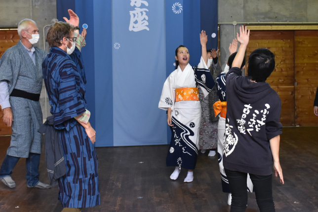A group of people dressed in traditional Japanese clothing, engaging in a cultural dance or activity inside a wooden hall with a blue banner in the background.