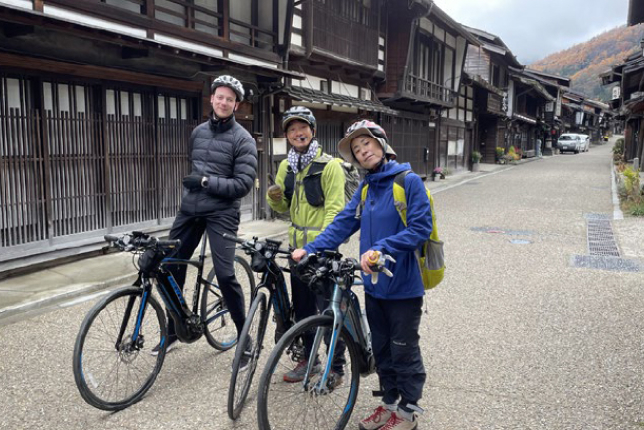 Three cyclists posing with their bikes in a historic Japanese town, surrounded by traditional wooden buildings.