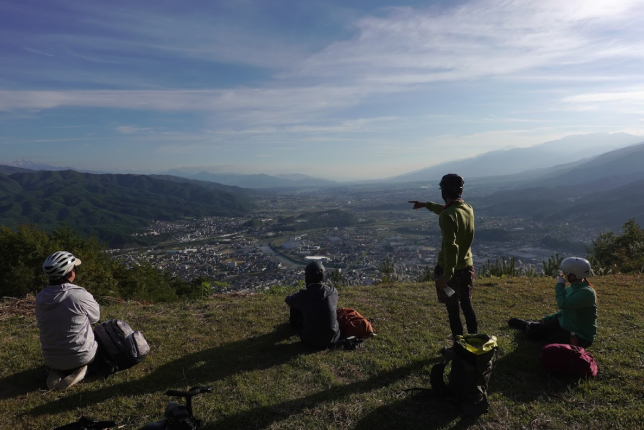 A group of adventurers sitting on a hilltop, overlooking a vast valley with a winding river, as one of them points toward the horizon.