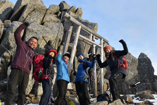 A group of hikers celebrate their ascent at the summit, standing in front of a wooden torii gate surrounded by large rocky formations.