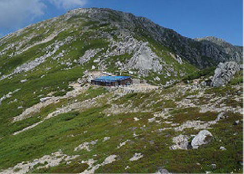 A mountain hut situated on a lush green slope with rocky terrain. The hut is surrounded by a scenic alpine landscape under a clear blue sky.
