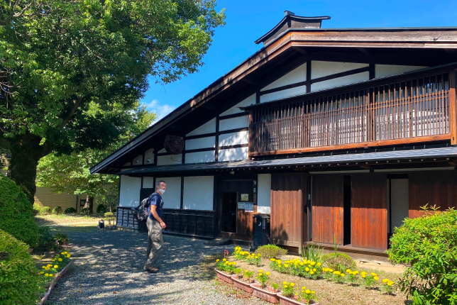 A traveler walking towards a traditional Japanese house with wooden architecture and a thatched roof, surrounded by greenery and yellow flowers on a sunny day.