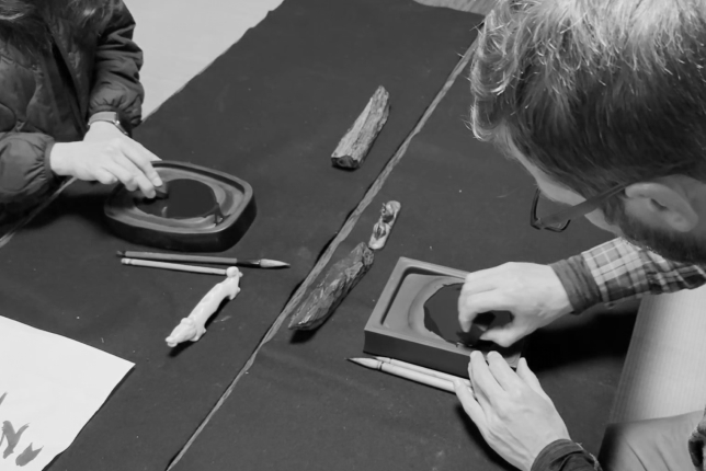 A man grinding an inkstick on an inkstone while another participant does the same, both preparing ink for calligraphy.