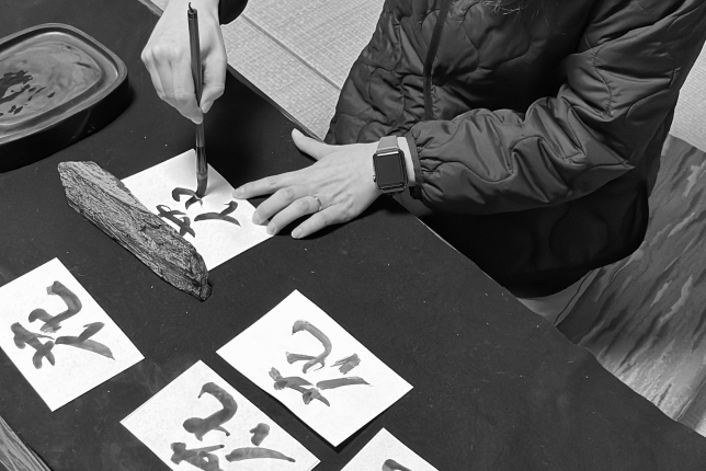 A close-up of a person writing Japanese calligraphy on paper using a traditional brush, with multiple completed calligraphy sheets visible.