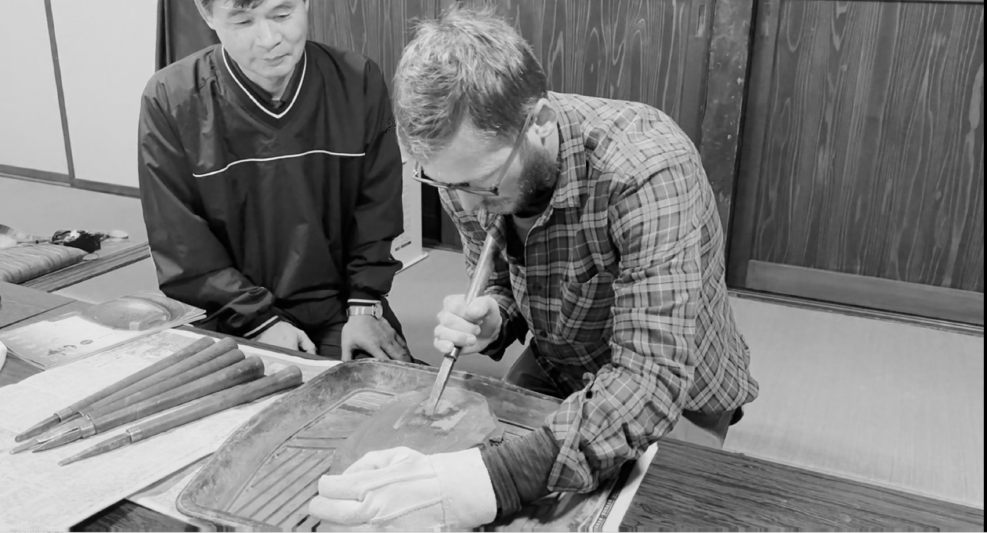 A craftsman demonstrates traditional wood carving techniques to a visitor in a Japanese-style room.