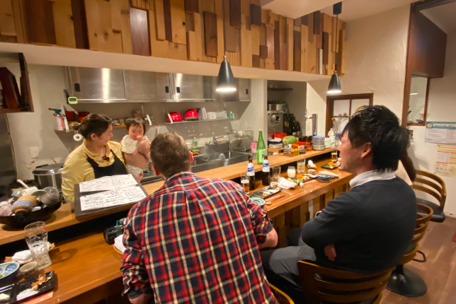 Two customers enjoy conversation and drinks at a cozy Japanese izakaya counter while the chef prepares food.