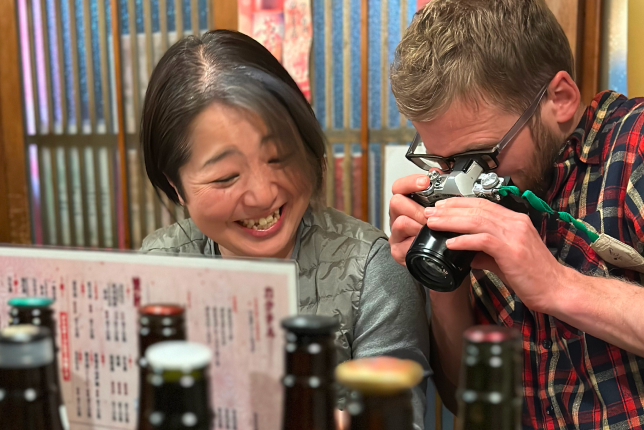 A smiling local woman and a foreign traveler share a laugh as he takes a close-up photo of the menu at a Japanese sake bar.
