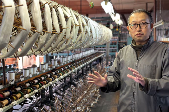A guide explaining the traditional silk spinning process in a factory filled with spinning reels and silk threads. Rows of machinery showcase the intricate process of silk production.