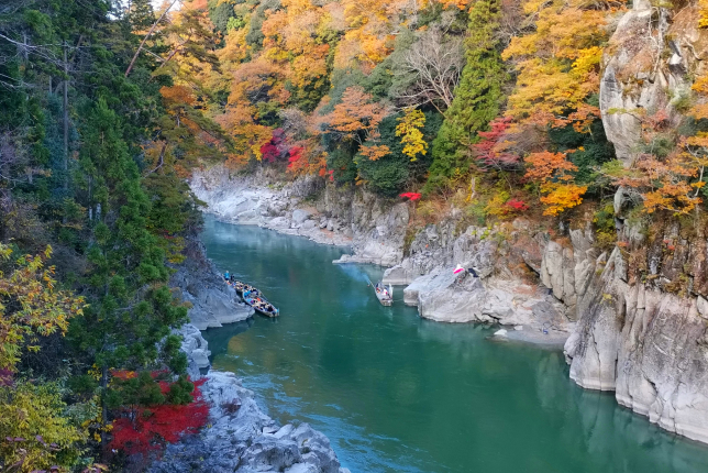 A picturesque river canyon during autumn, with vibrant red, orange, and yellow leaves reflecting on the calm water, as boats navigate the scenic landscape.