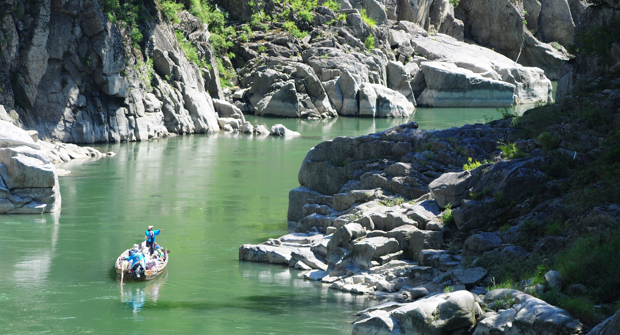 A traditional wooden boat gliding through a green river surrounded by large rock formations under bright sunlight.