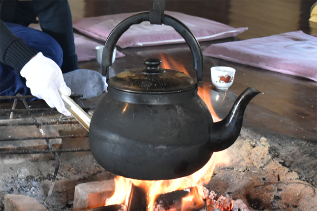 A black iron kettle hanging over an open fire in a traditional Japanese hearth, with hands adjusting its handle.