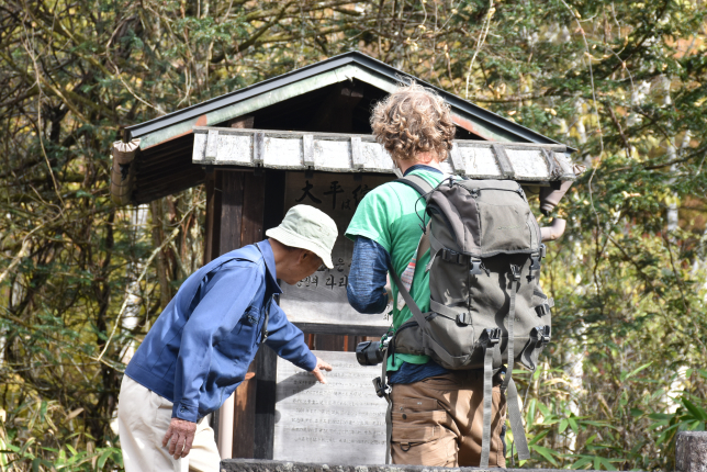 An elderly Japanese man pointing at an information board while explaining something to a foreign traveler wearing a backpack, surrounded by nature.