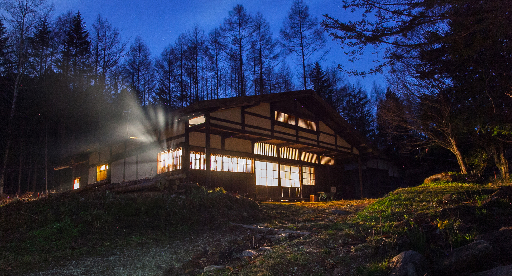 A traditional Japanese house in a forest, illuminated warmly at dusk, with trees silhouetted against the darkening sky.