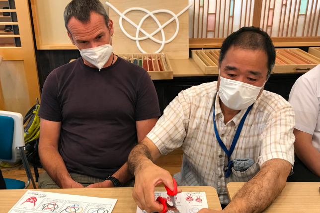A Japanese instructor demonstrating the art of creating mizuhiki (decorative paper cord) knots, as a foreign participant observes attentively.