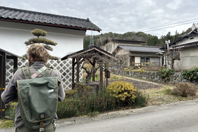 A traveler with a green backpack walking through a Japanese village with traditional architecture and neatly arranged gardens.
