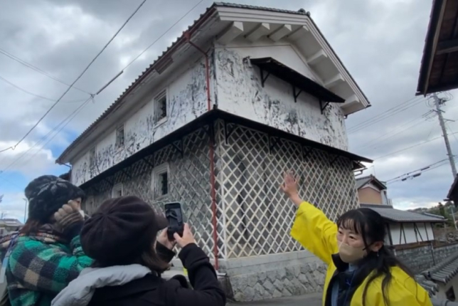 A tour guide in a yellow jacket enthusiastically explaining the history of a historic Japanese warehouse with a white plastered and lattice-patterned wall, as visitors take photos.