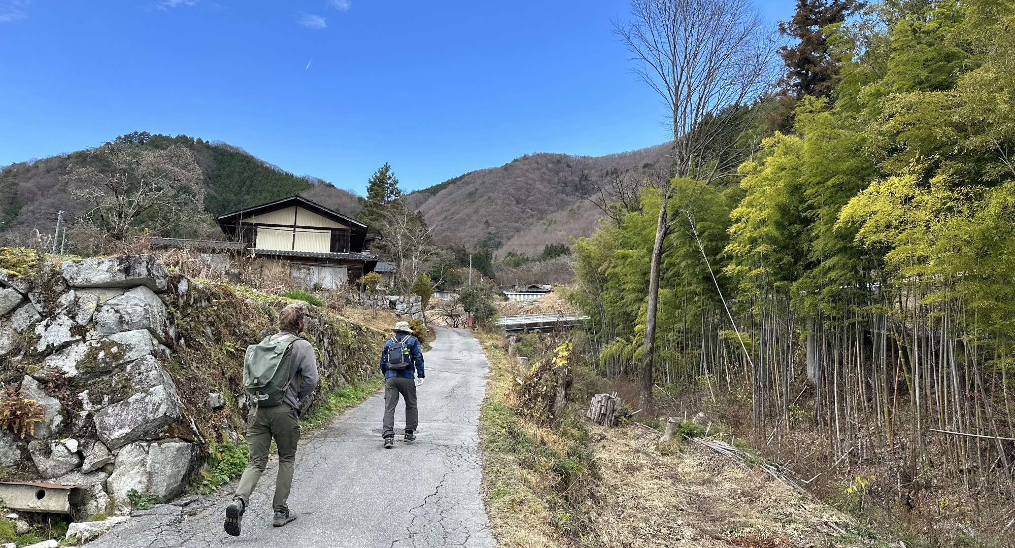Two people hiking on a rural path surrounded by mountains, bamboo forests, and traditional Japanese houses under a bright blue sky.