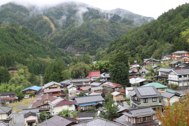A scenic view of a small mountain village surrounded by lush green forests and mist-covered hills.