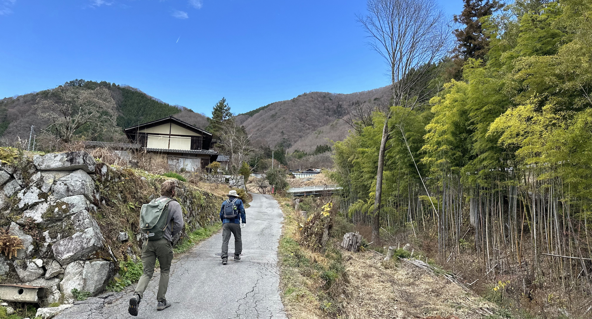 Two people hiking on a rural path surrounded by mountains, bamboo forests, and traditional Japanese houses under a bright blue sky.