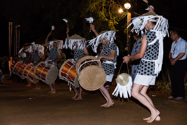 A group of taiko drummers in matching black-and-white outfits with decorative headpieces perform at night, energetically raising their drumsticks while playing large traditional drums.