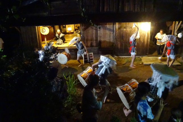 A vibrant nighttime scene of a Japanese festival with drummers and dancers performing in front of an old wooden house, illuminated by warm lighting. The performers are in motion, holding their drums and raising their hands.