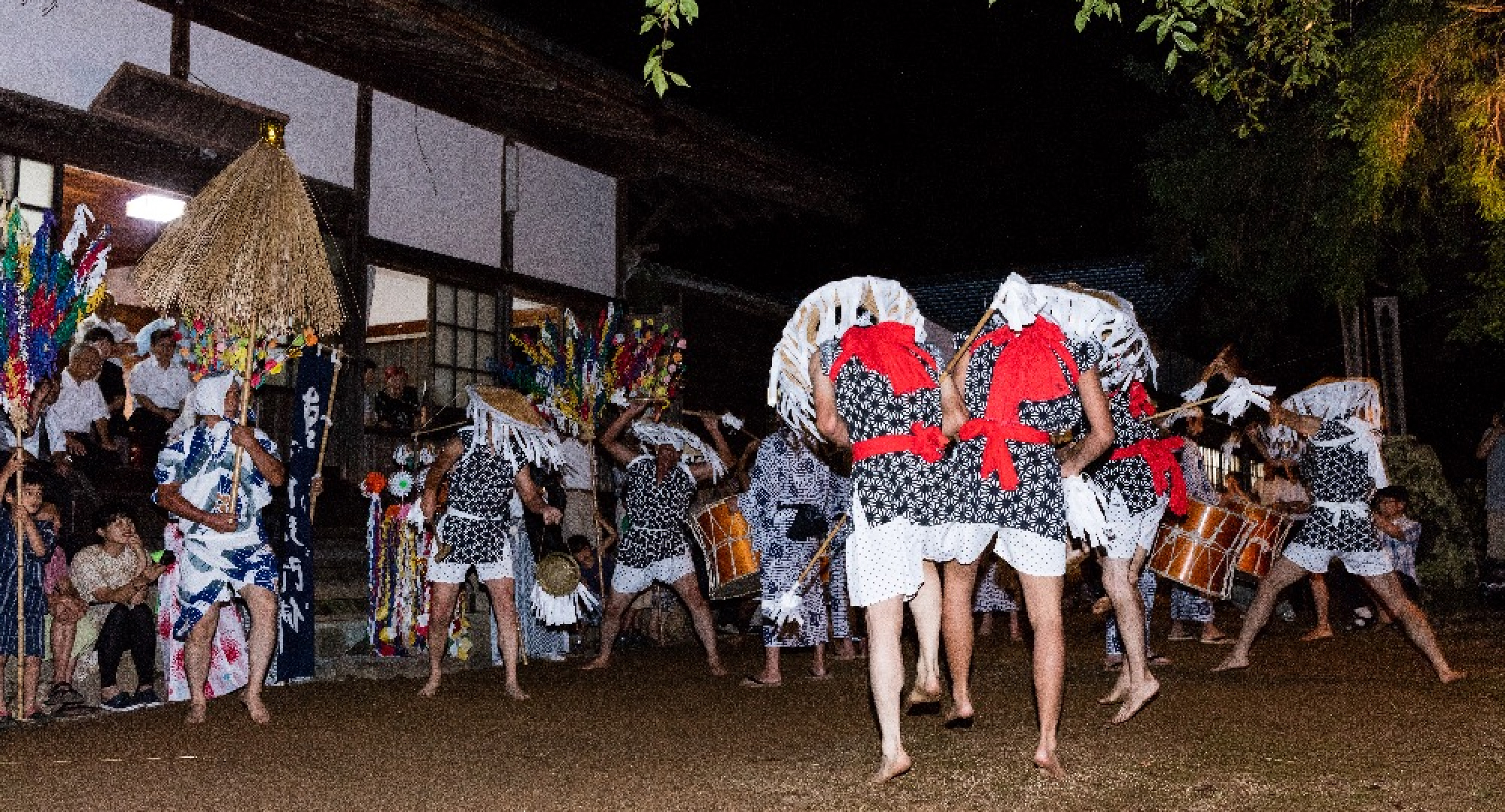 A nighttime traditional Japanese festival featuring performers in black-and-white patterned outfits with red sashes, playing large taiko drums and dancing in a lively atmosphere. Spectators watch from the sidelines.