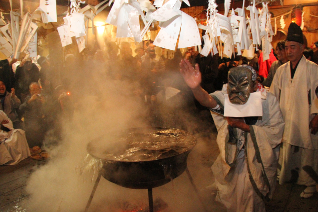 A sacred ritual at a Shinto shrine, where a person wearing a mask and white robes performs a ceremony over a large steaming cauldron, surrounded by an audience.