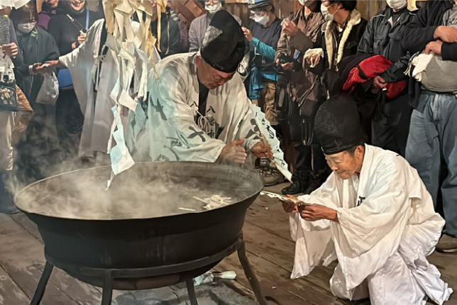 Two Shinto priests dressed in white robes performing a purification ritual, stirring a steaming cauldron while being observed by a crowd.