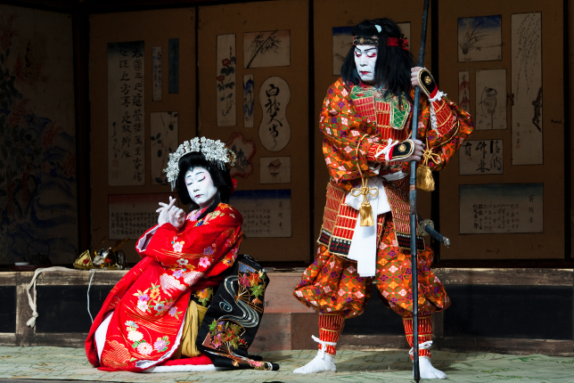 A kabuki performance featuring two actors in ornate costumes—one dressed as a samurai and the other in a red kimono—engaged in an intense scene.