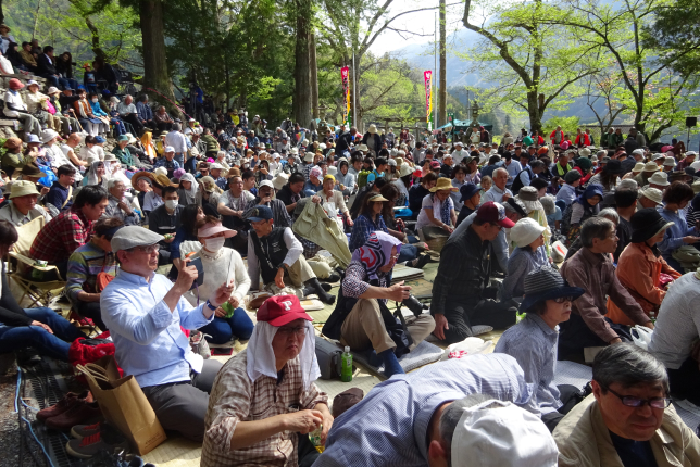 A large audience gathered outdoors, sitting on mats and watching a traditional performance, dressed in casual and traditional clothing.