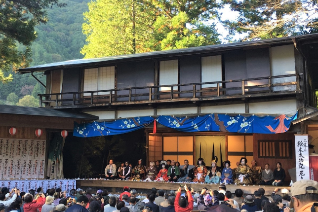 An open-air stage set against a scenic backdrop, where performers in kabuki costumes entertain a large audience.