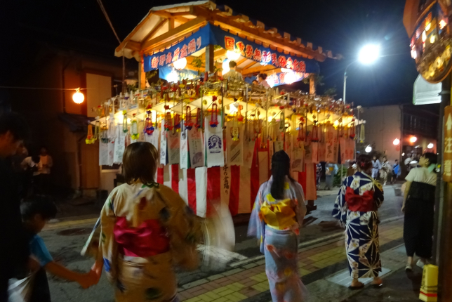 Women in colorful yukata walking and dancing near a beautifully decorated festival float adorned with paper lanterns and banners.