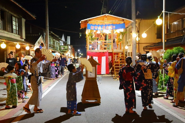 A nighttime festival scene with children and adults in yukata dancing along a street lined with glowing lanterns and a raised festival stage.