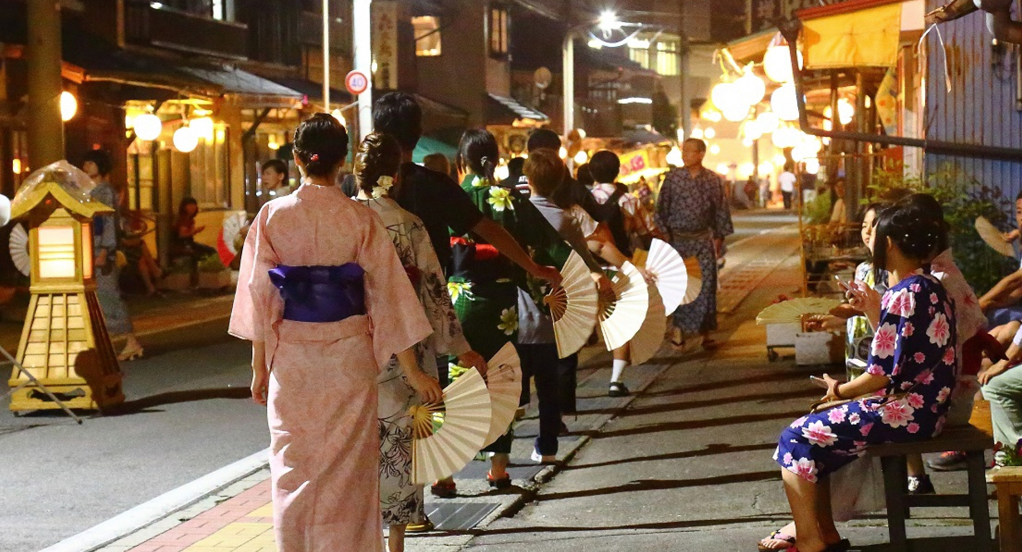 A group of people wearing yukata (summer kimono) dancing gracefully with fans during a traditional Japanese festival at night, illuminated by warm lantern lights.