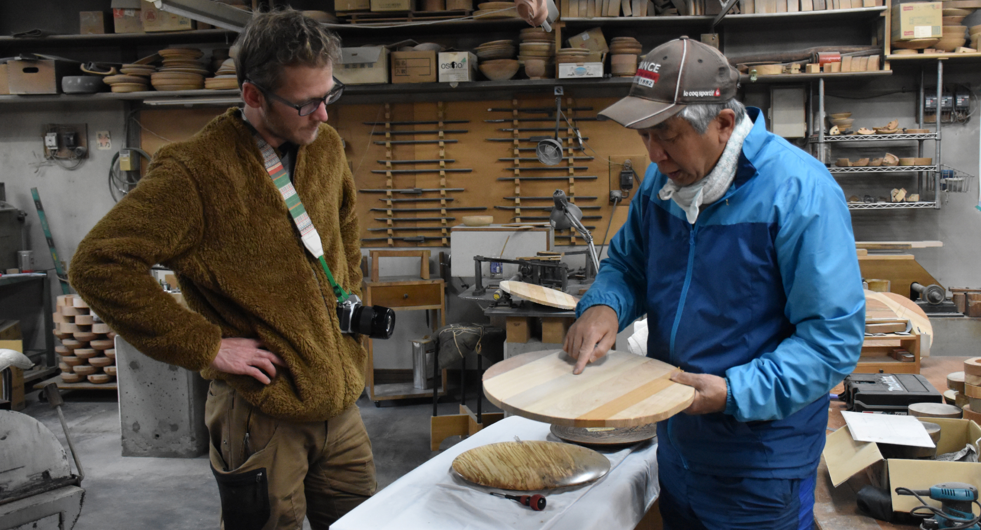 A master craftsman in a blue jacket explaining the details of a wooden piece to a visitor in a woodworking studio. The background is filled with woodworking tools and materials.