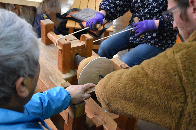 Participants working on a wooden plate using a traditional lathe, guided by an experienced craftsman. The workshop setting highlights the hands-on learning experience.